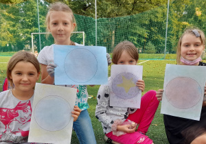 Cztery uśmiechnięte dziewczynki siedzą na boisku szkolnym i pokazują zaprojektowane przez siebie z apomocą kredy duże kropki. Jedna z dziewczynek zakrywa sobie część twarzy swoją pracą.
